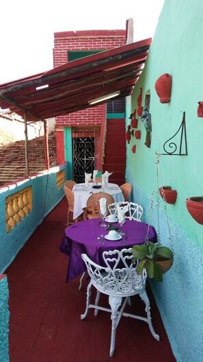 A small outdoor dining area with a table set for two, surrounded by white chairs, on a balcony with a red floor and a blue wall. - Caridad and Friends Rooms (Trinidad)