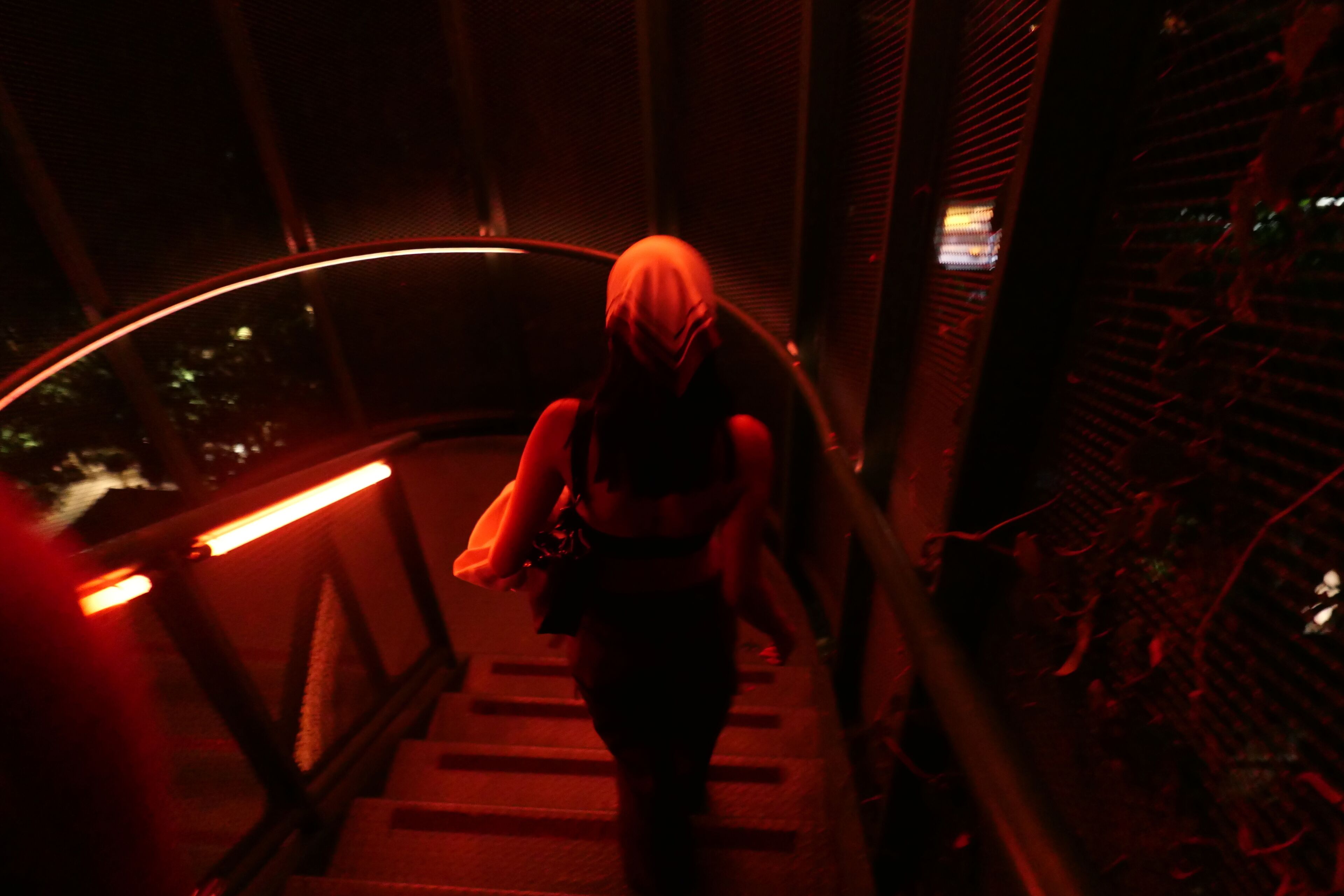 Una persona baja una escalera en un área poco iluminada, con barandales de metal y vista al exterior.