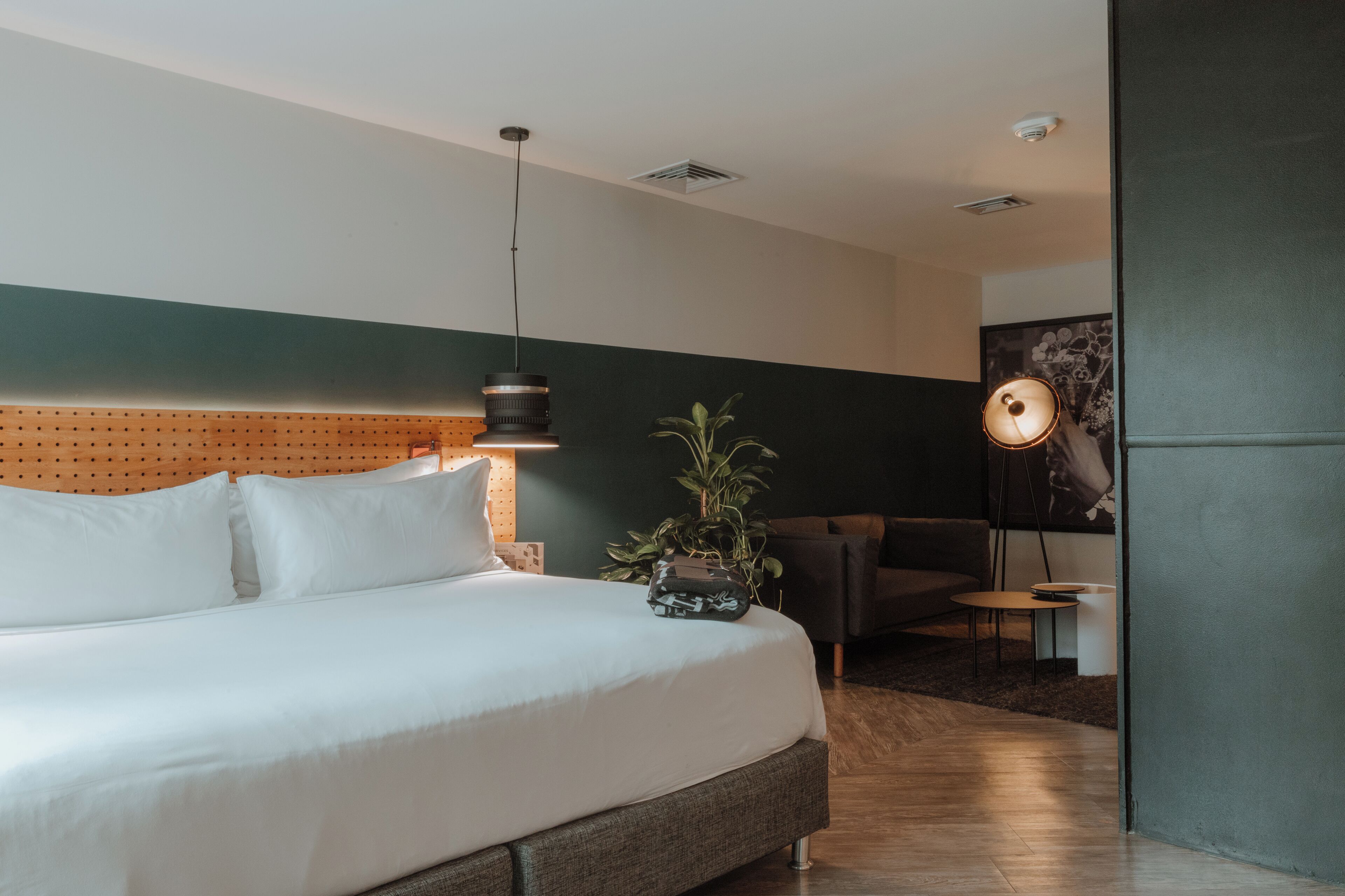 Une chambre d’hôtel moderne avec un grand lit, une tête de lit en bois, une plante en pot, une table d’appoint et une petite table ronde.