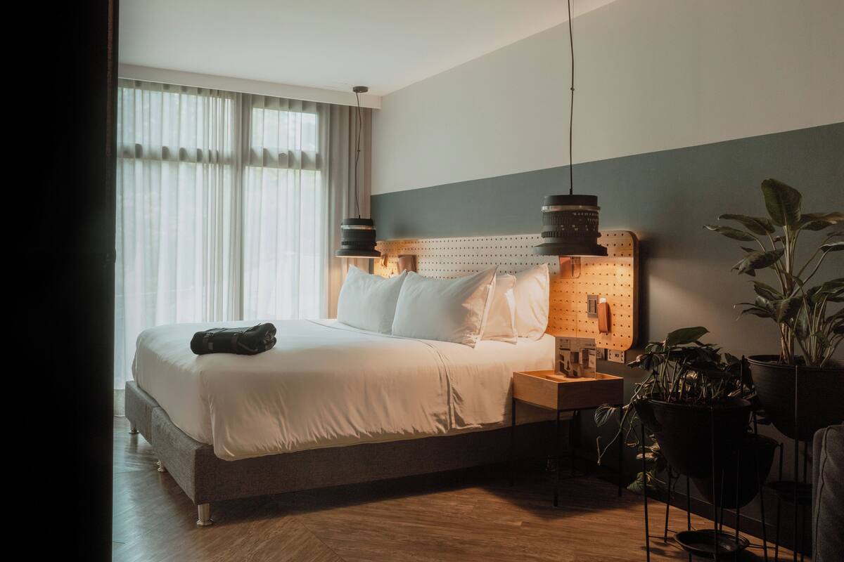 A neatly made bed with pillows and a blanket, a bedside table with a lamp, and a wall-mounted phone.