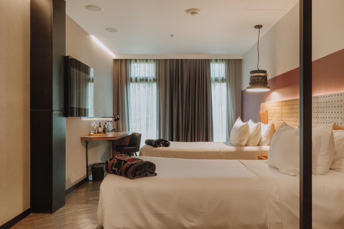 A hotel room with two beds, a desk, a chair, a lamp, and a large window with curtains.