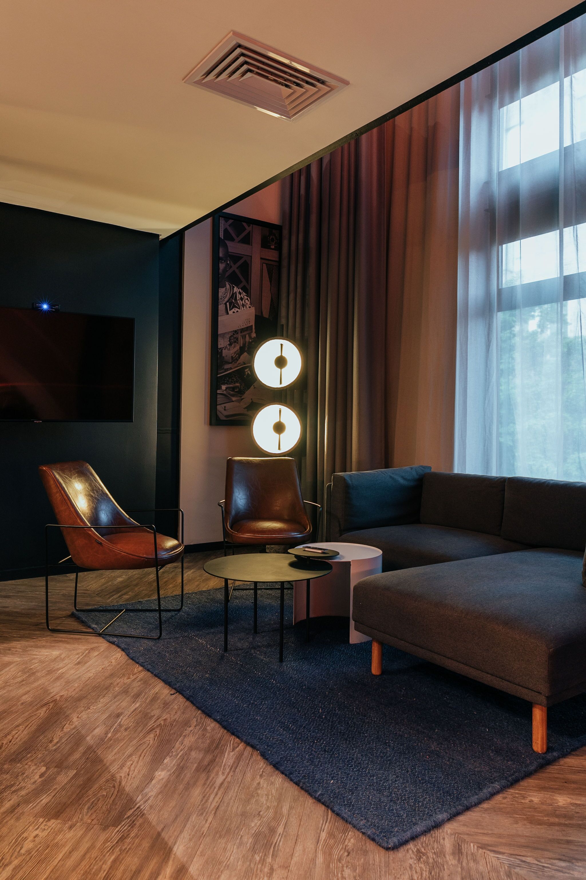 A modern living room with a dark-colored sofa, two leather chairs, a small round table, and a flat-screen TV mounted on the wall.