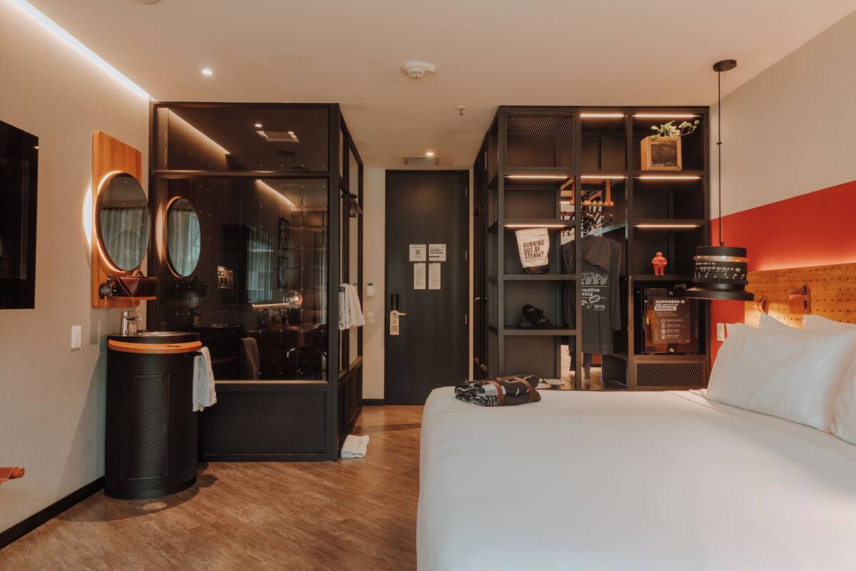 A modern hotel room with a large bed, a vanity with a mirror, a small bathroom, and a built-in bookshelf.