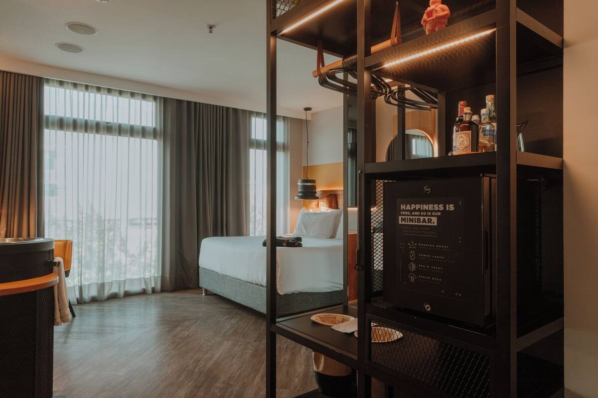 A hotel room with a bed, a desk, a minibar, and a large window with curtains.