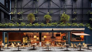 Un café al aire libre con mesas y sillas, una zona de bar y un edificio con fachada oscura y vegetación en el fondo.