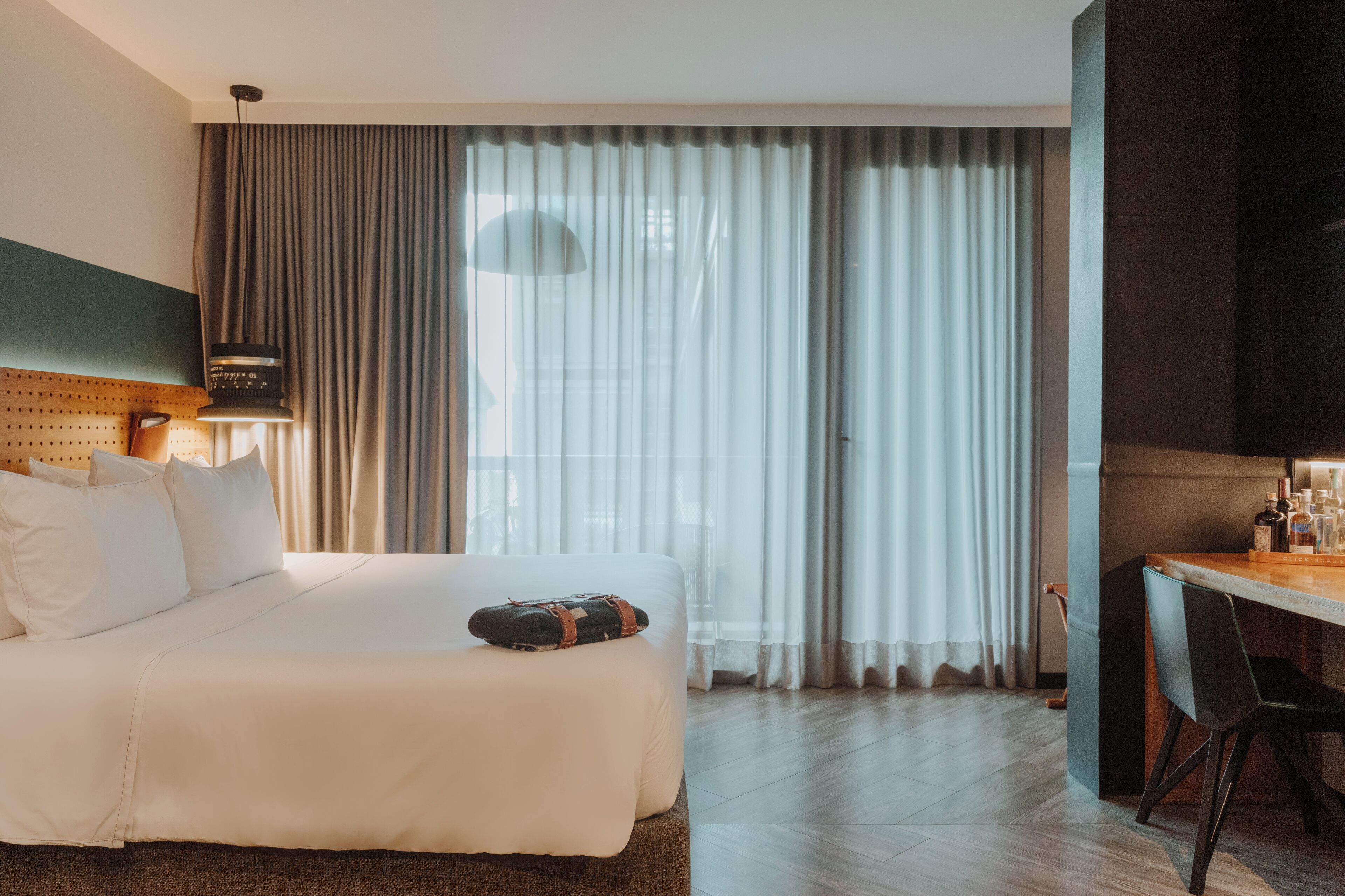 Une chambre d’hôtel avec un grand lit, un bureau avec une chaise, une lampe et une vue à travers des rideaux transparents.