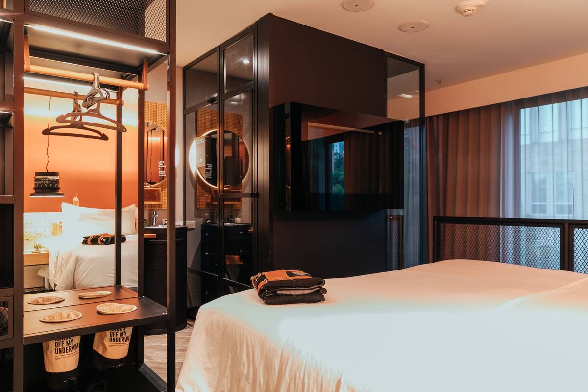A modern hotel room with a large bed, a TV mounted on the wall, and a mirror above the bed.