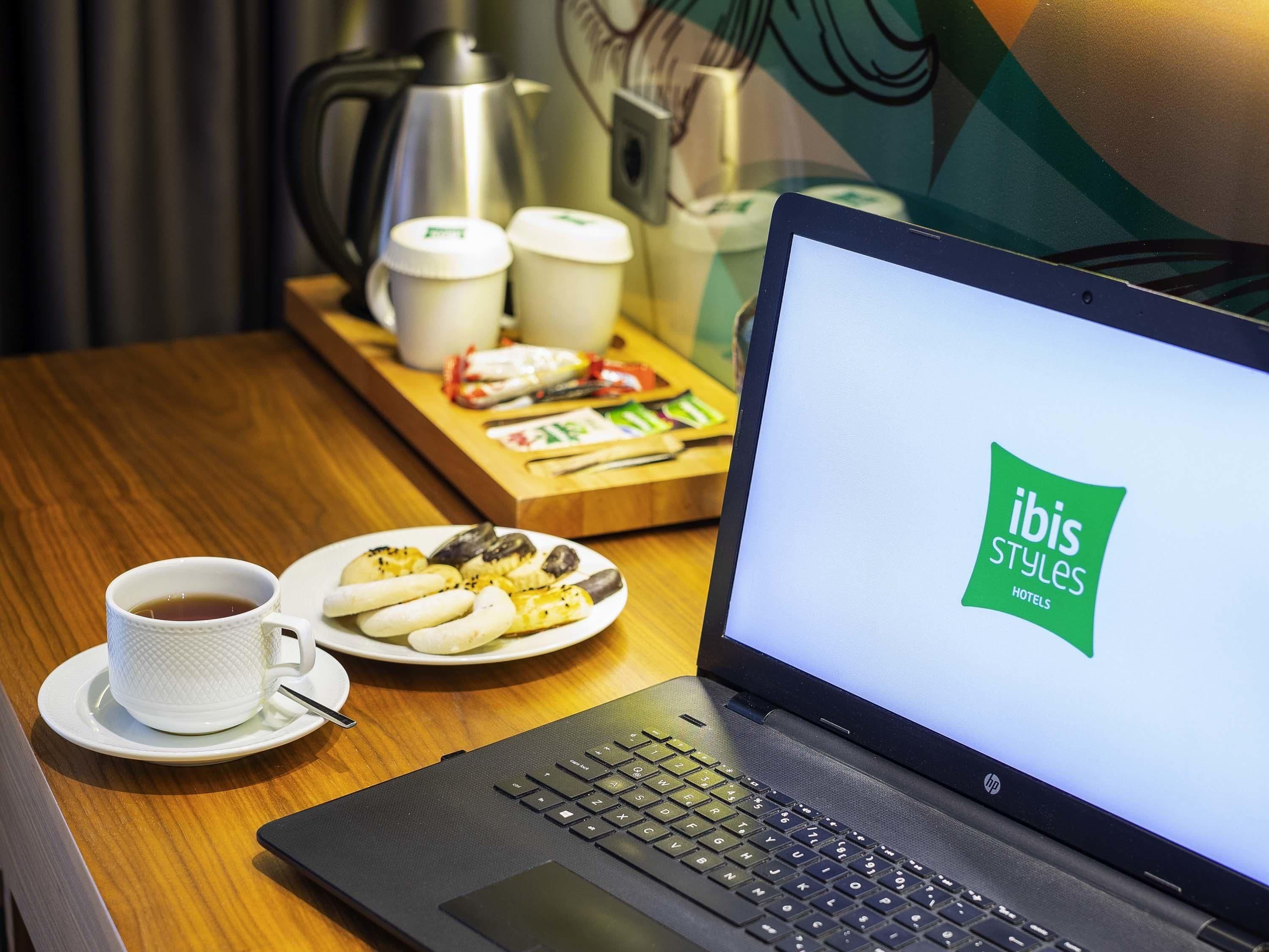 A laptop on a wooden table with a cup of tea, a plate of food, and a coffee maker in the background.