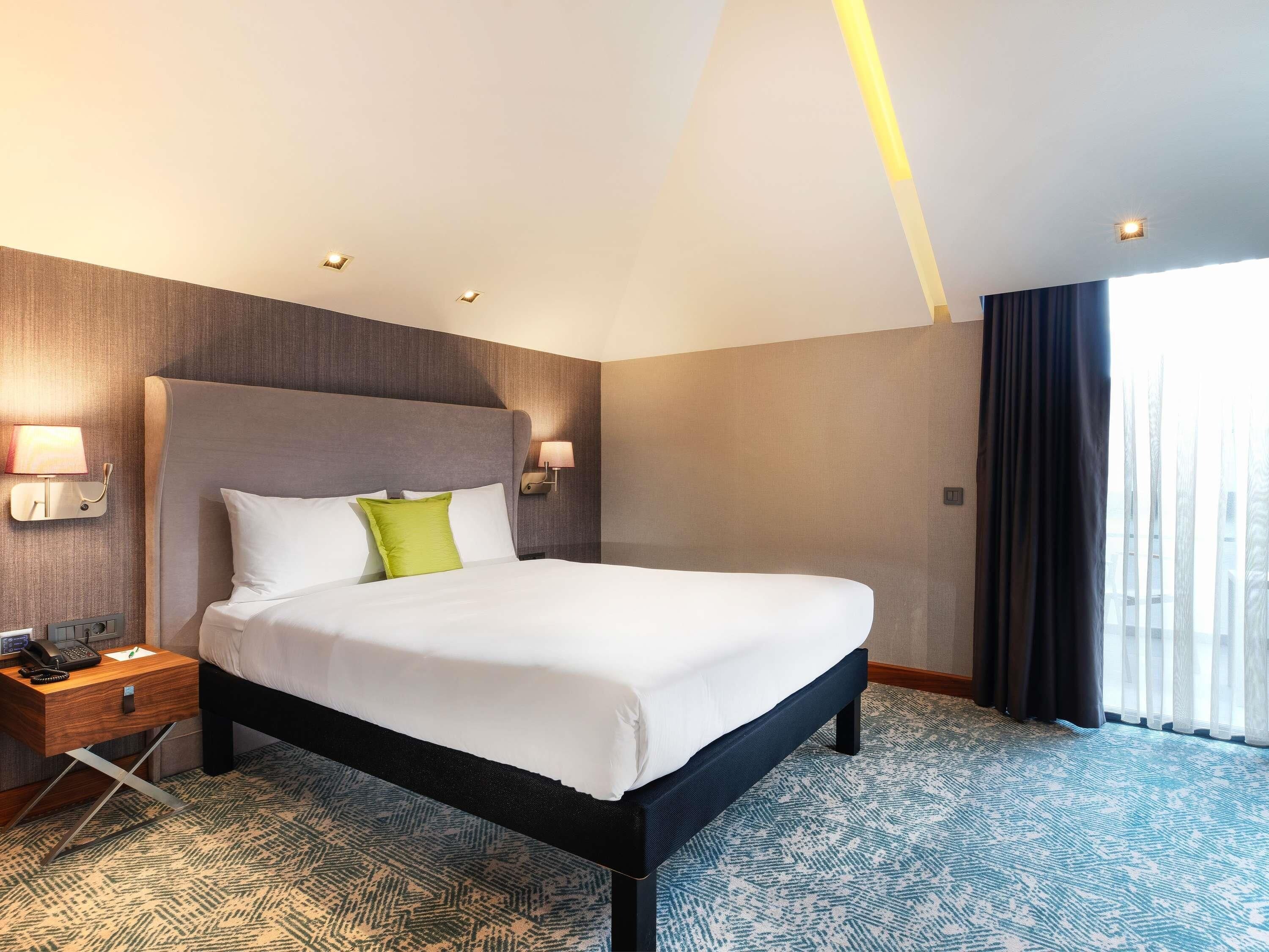A hotel room with a large bed, bedside lamps, a nightstand, and a view of the outside through a window.