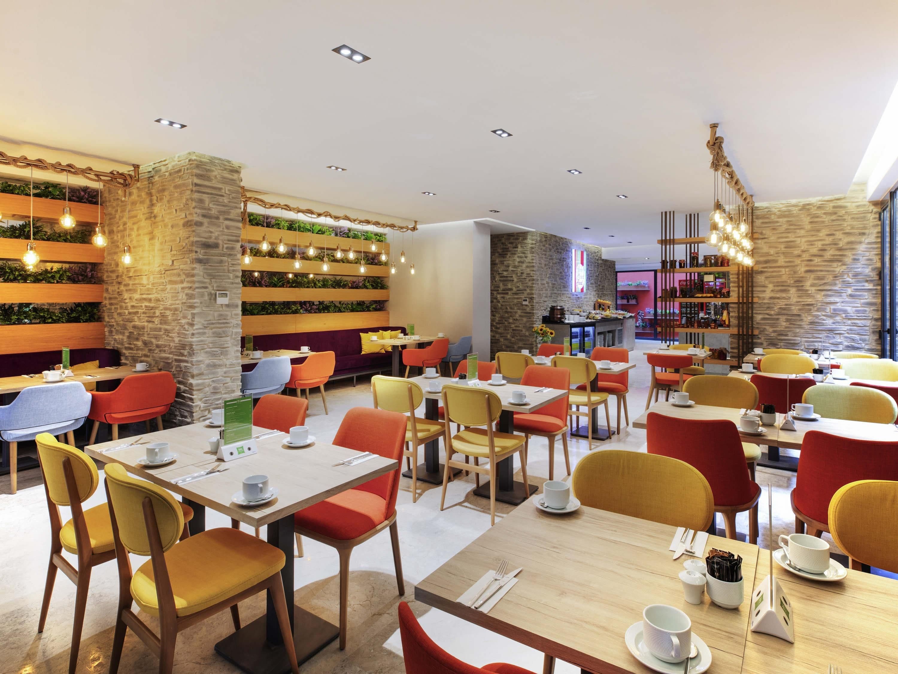 A modern restaurant with wooden tables and chairs, a stone wall with greenery, and a bar area.
