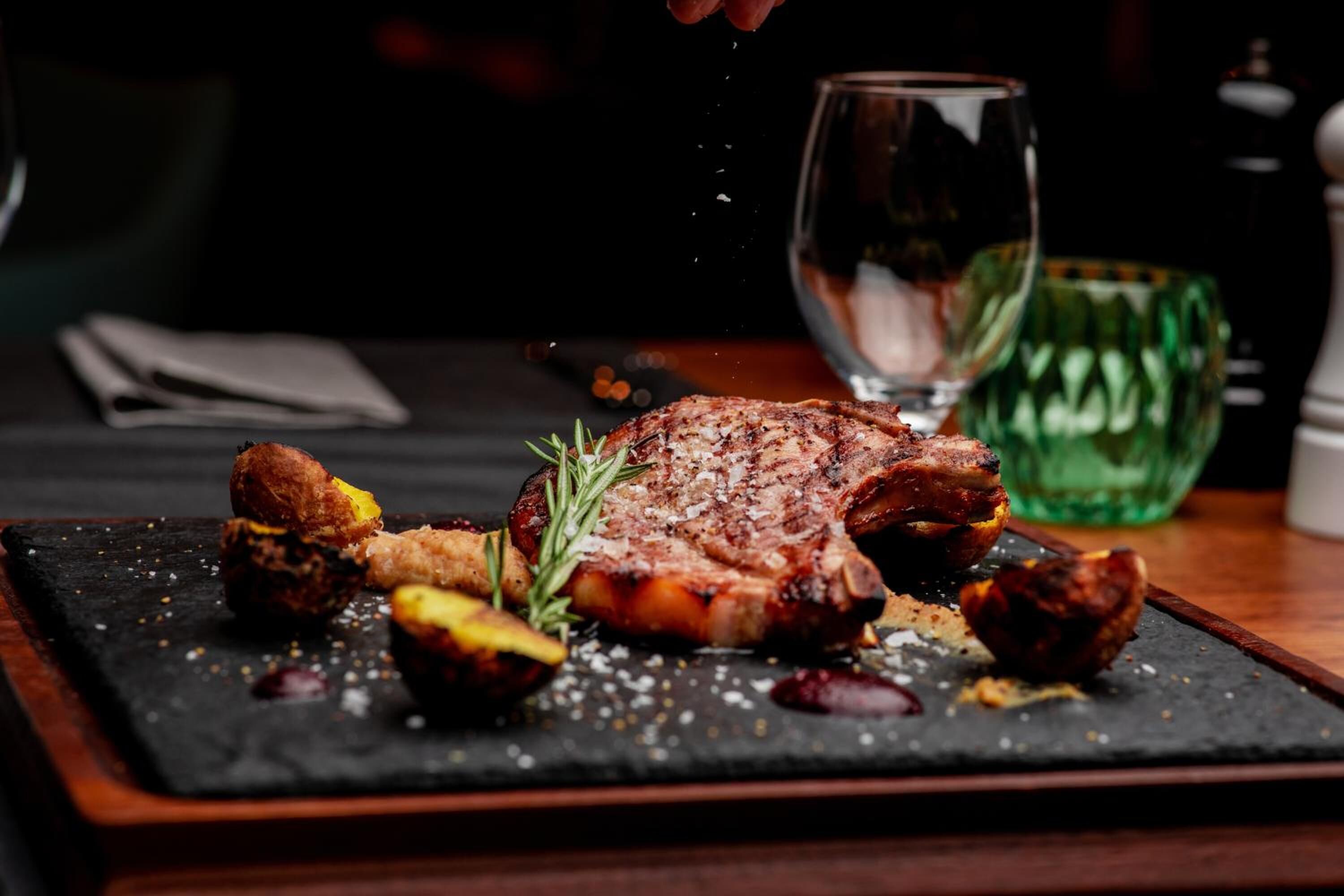 A steak with rosemary and potatoes on a slate plate, a glass of water, and a green glass in the background.