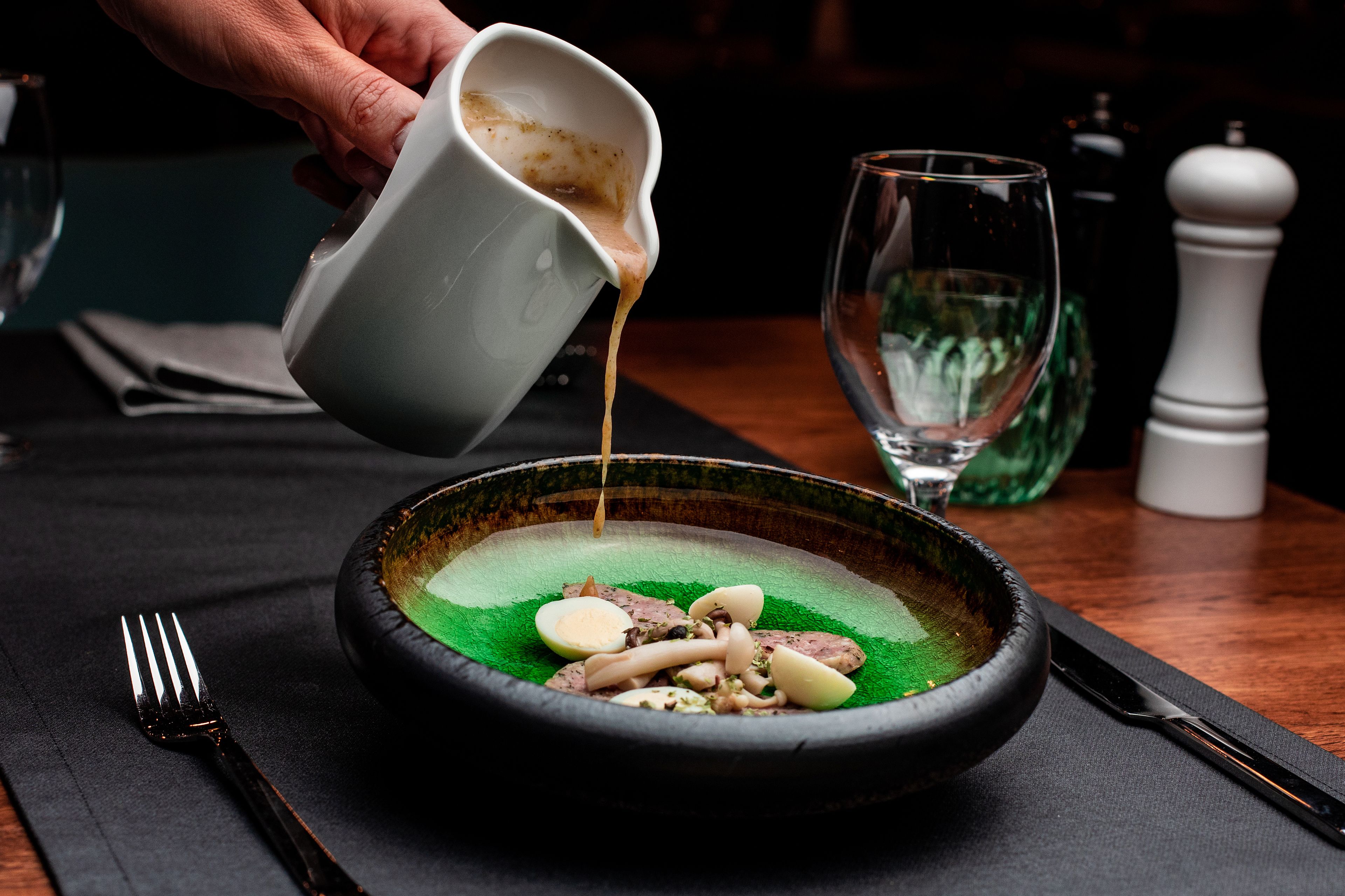 A bowl of food with a sauce being poured over it, a fork, and a knife on a table.