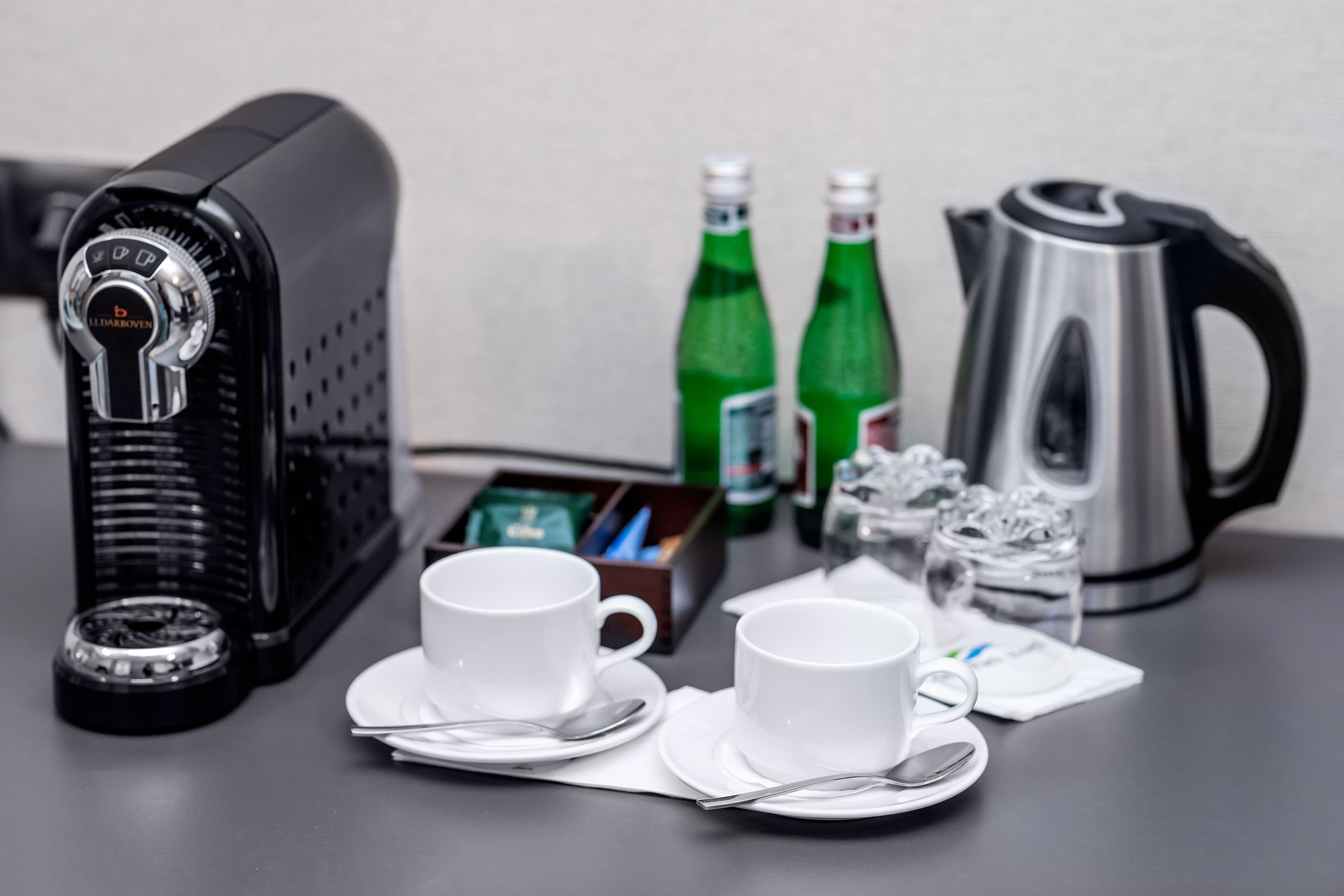 Ein Tisch mit Kaffeemaschine, zwei Tassen, einem Wasserkocher und zwei Wasserflaschen.