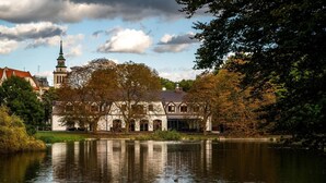 Un edificio histórico con una torre de reloj, ubicado junto a un lago tranquilo, rodeado de árboles y otros edificios.