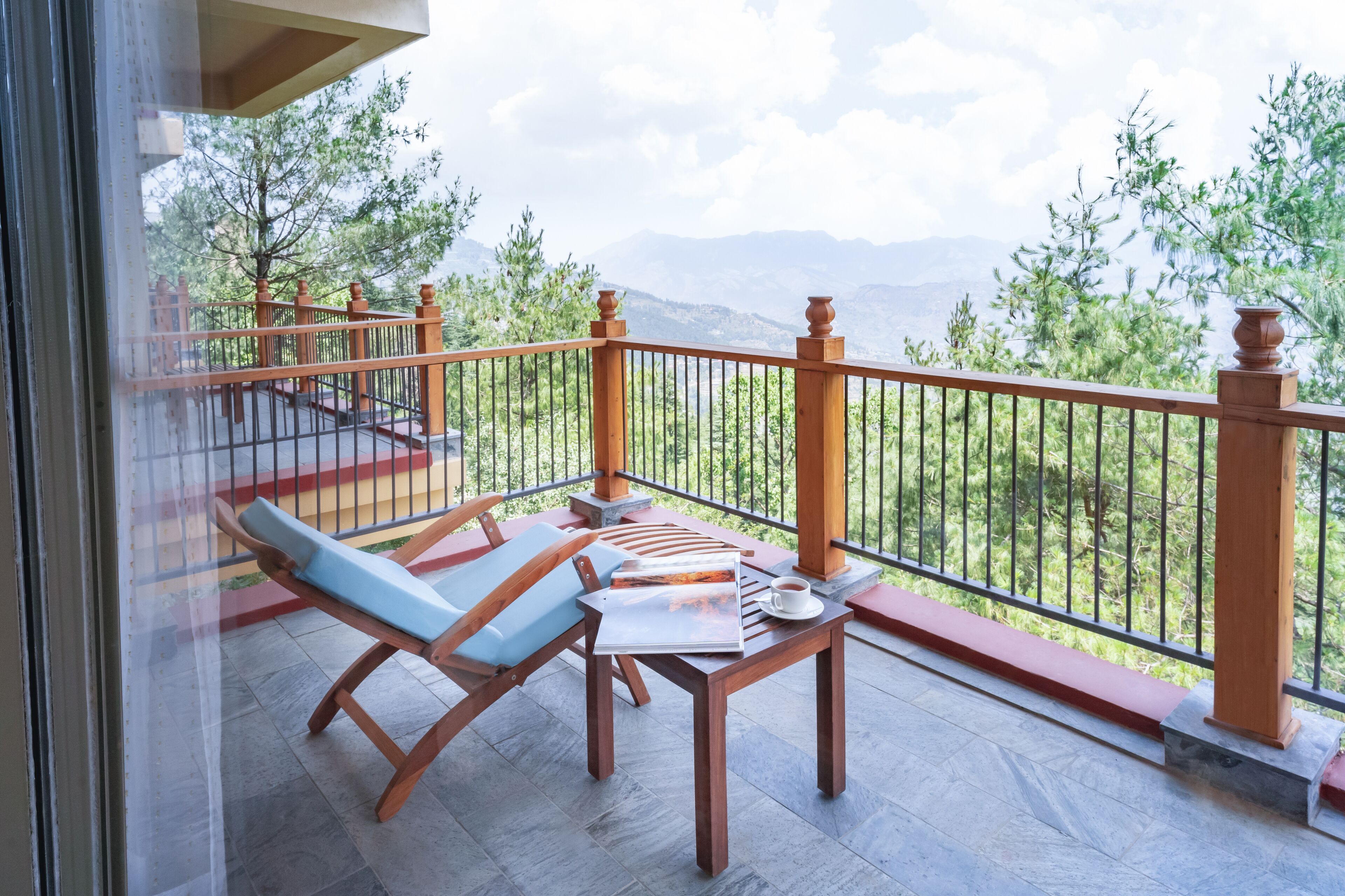 A balcony with a wooden table, a blue cushioned chair, and a view of mountains and trees.