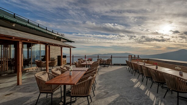 An outdoor terrace with wooden tables and chairs, a glass wall, and a view of mountains. - Taj Theog Resort & Spa, Shimla (Theog)