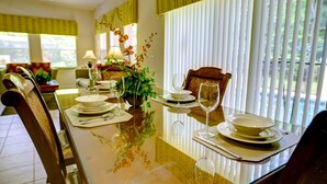 A dining table set for four with chairs, a vase with flowers, and a view of the outdoors through large windows. - Durham Townhome with Pool at Encantada (Kissimmee)