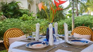 Une table dressée pour un repas en plein air, avec des chaises en osier, une nappe blanche et un vase rempli de fleurs rouges.