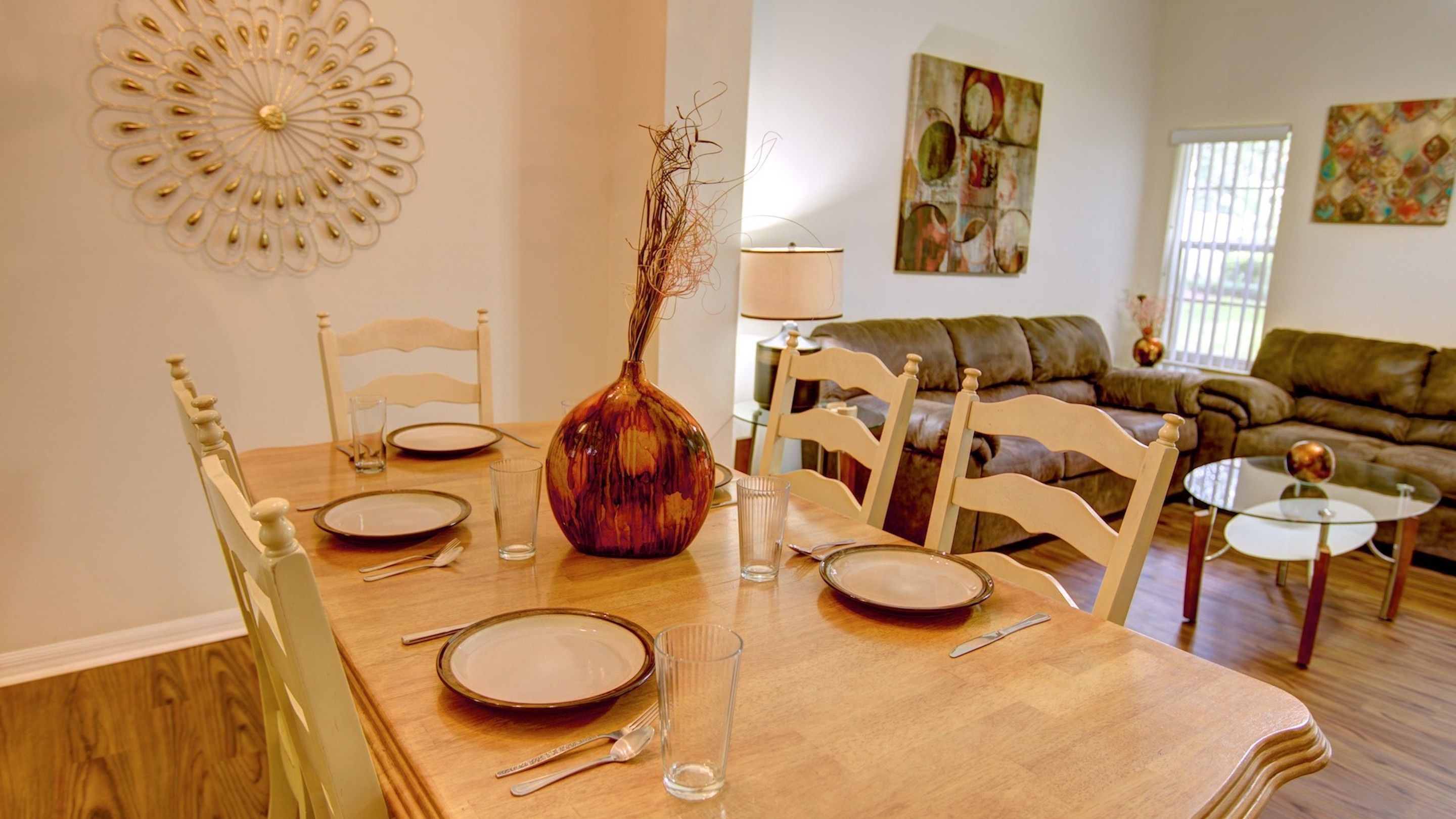 Un comedor con una mesa de madera dispuesta para cuatro personas, rodeada de sillas de madera. Hay un jarrón con una rama sobre la mesa, y un vaso de agua. En el fondo, se ve un sofá y una mesa baja.