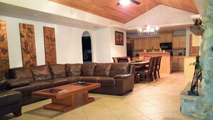 Amplia sala de estar con sofá de cuero, mesa de centro de madera y una mesa de comedor. Se ve una cocina al fondo con armarios de madera y un muro con detalles de piedra.