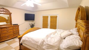 Un dormitorio con un aparador de madera, una cama con ropa de cama blanca, un televisor montado en la pared y una ventana con cortinas amarillas.