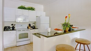 A modern kitchen with white cabinets, a built-in oven, a microwave, and a refrigerator. There is a small island with a plant and a cup.