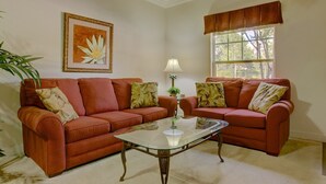 A living room with two red sofas, a glass coffee table, a framed floral artwork, a window with a view of trees, and a potted plant.