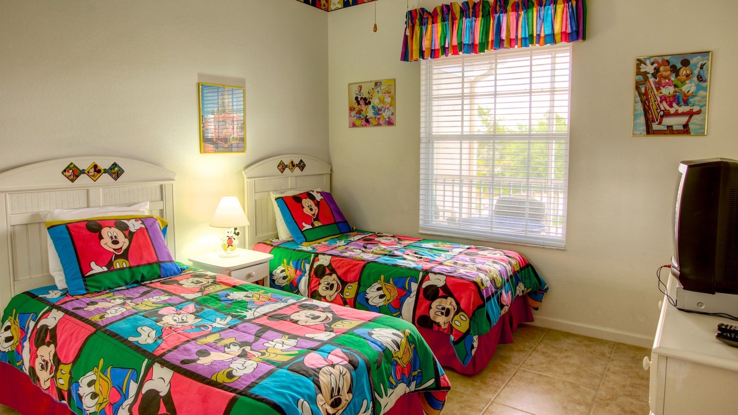 Ein Schlafzimmer mit zwei Betten, beide mit einem Bettbezug im Mickey-Mouse-Design. Dazwischen steht ein Nachttisch mit einer Lampe, und rechts daneben ein Fernseher auf einem Standfuß.