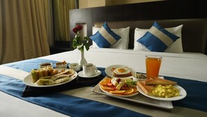 Ein ordentlich bezogenes Bett mit einer blauen und weißen Kissenanordnung, ein Tablett mit Frühstücksartikeln, darunter Toast, Eier und Obst, sowie eine Vase mit einer einzelnen roten Rose.