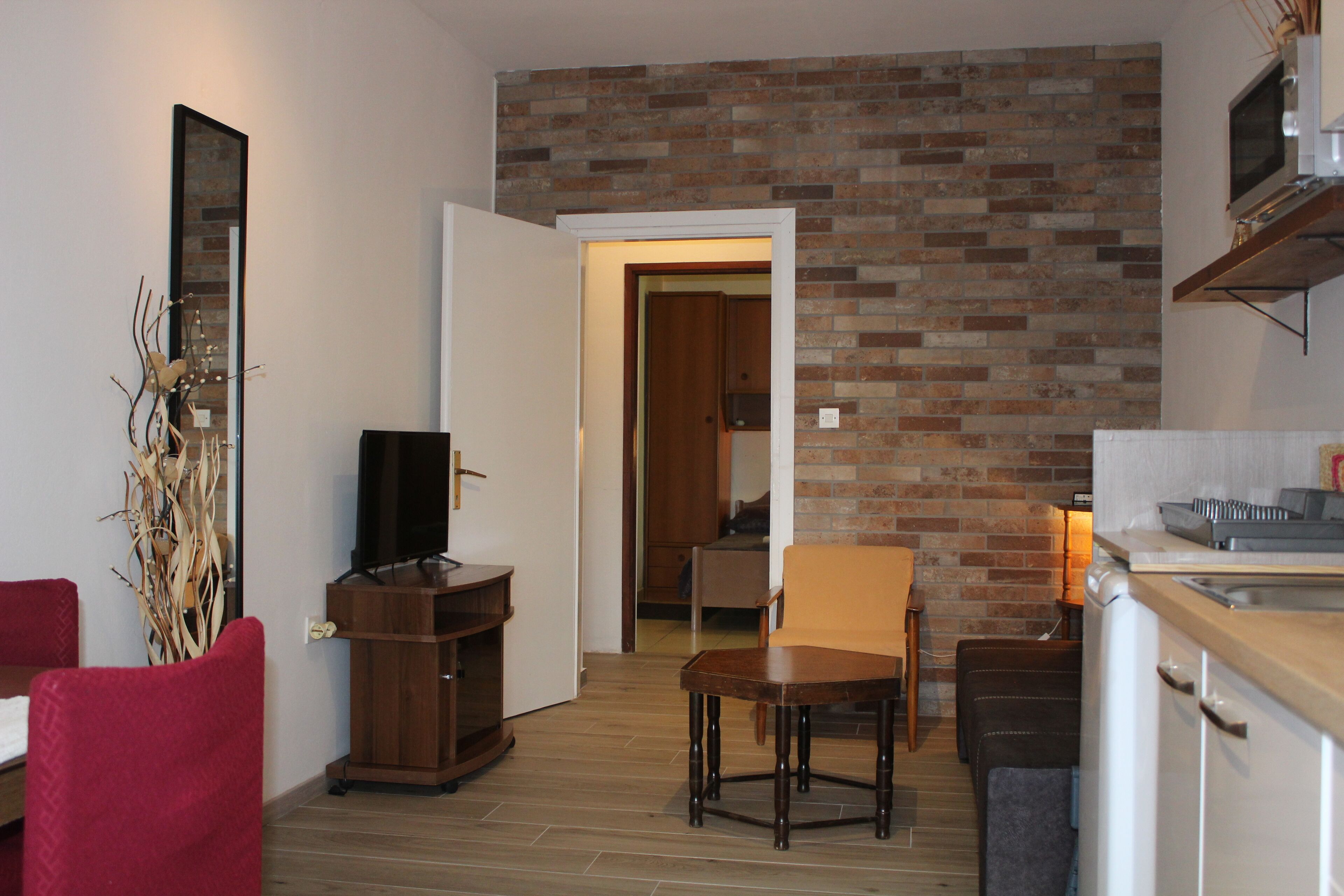 A compact living space with a brick wall, a television on a wooden stand, a red chair, a dining table with chairs, and a kitchen area with a microwave.