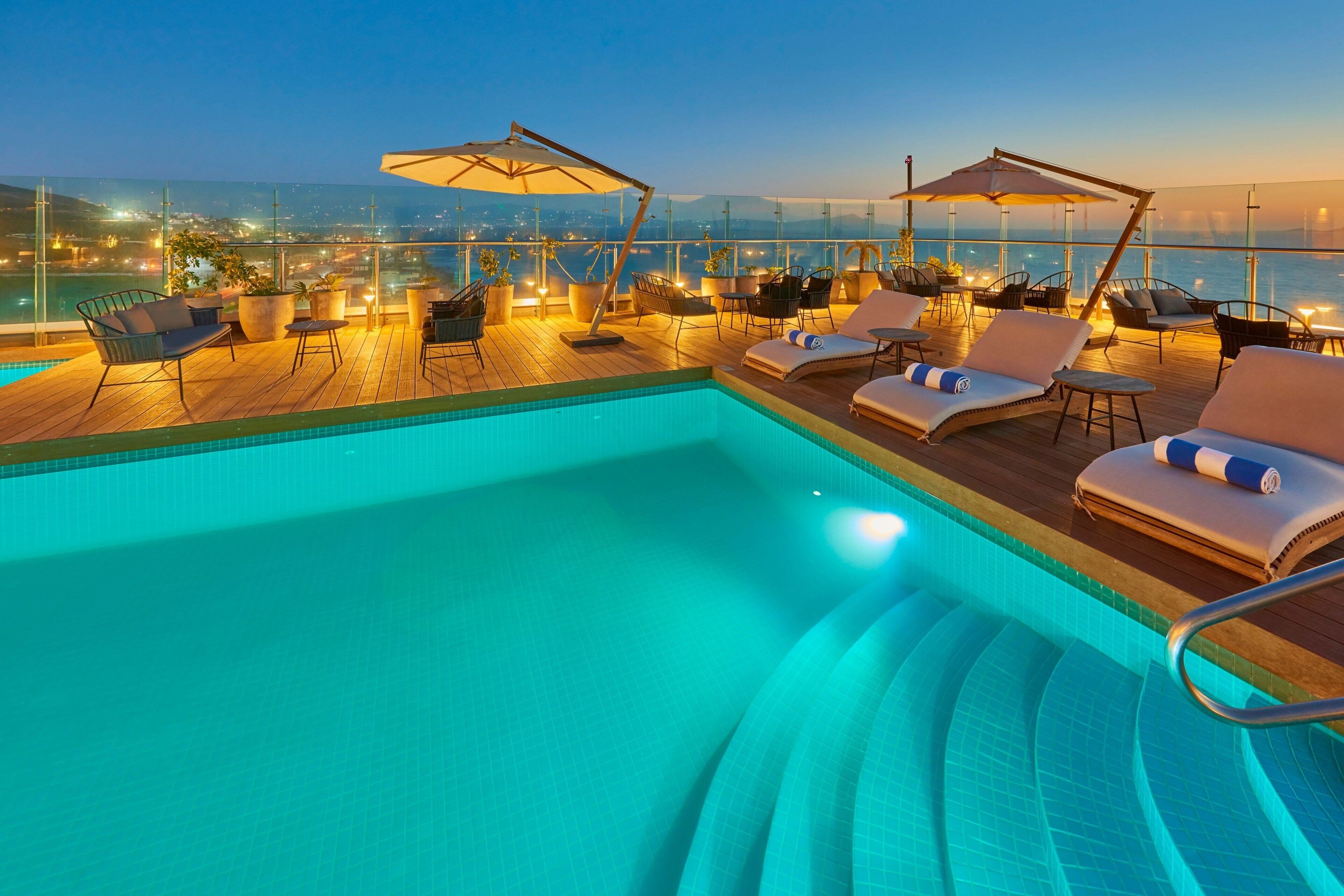 Un área de piscina en la azotea con sillones reclinables y sombrillas, con vista a un paisaje urbano al atardecer.
