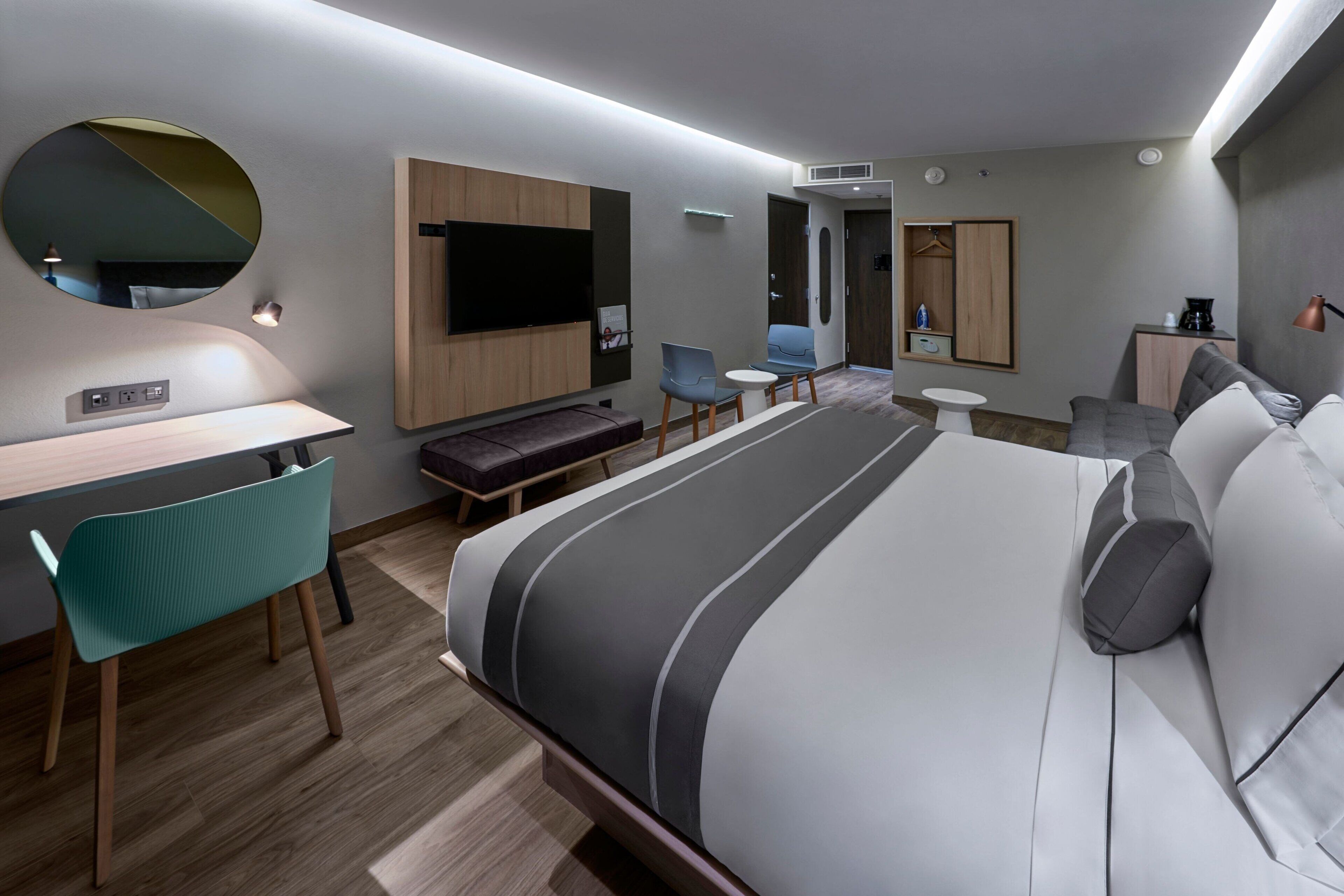 A modern hotel room with a large bed, a desk, a chair, a TV, and a small table.
