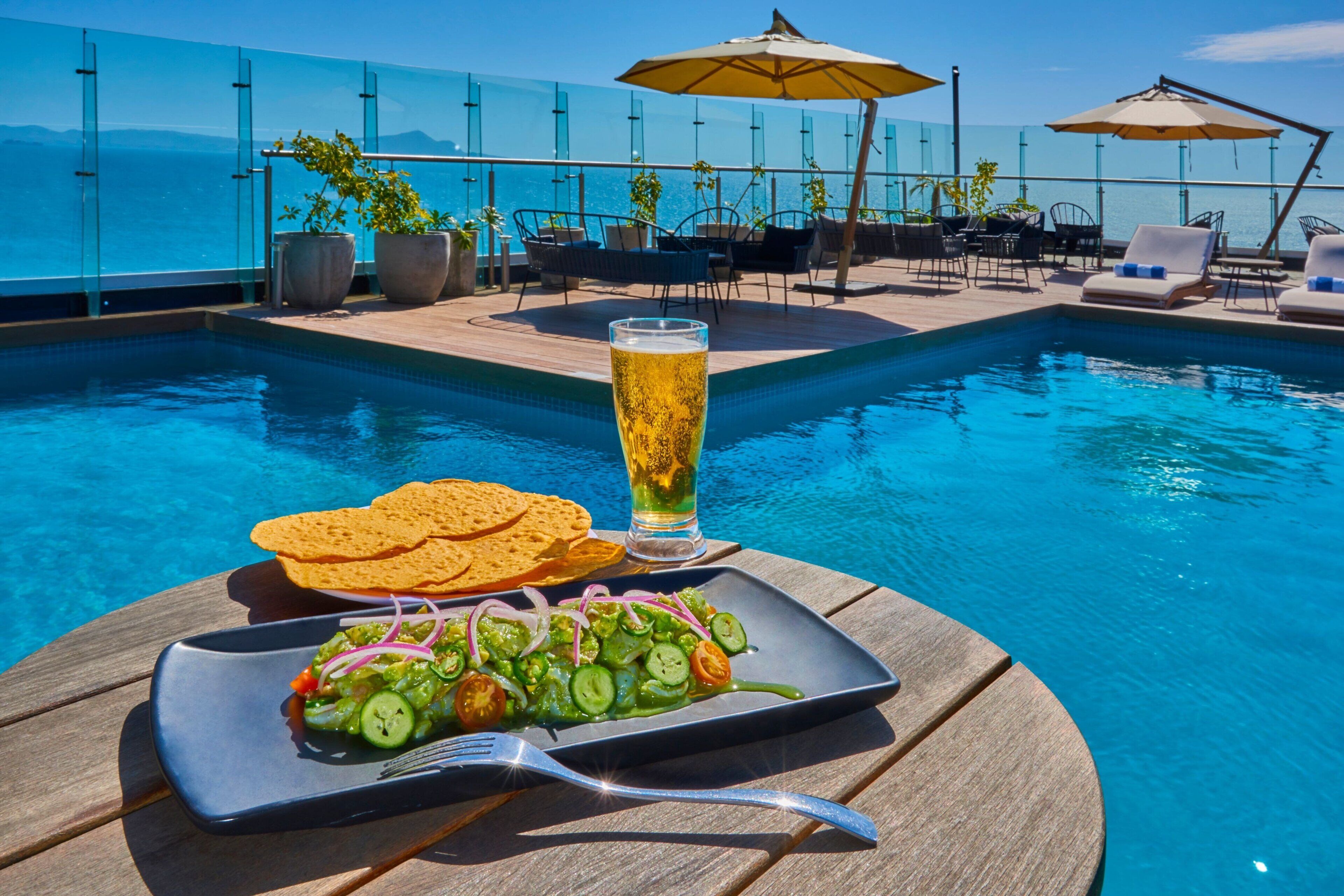Zona de comedor junto a la piscina con una mesa, una cerveza y un plato de comida.