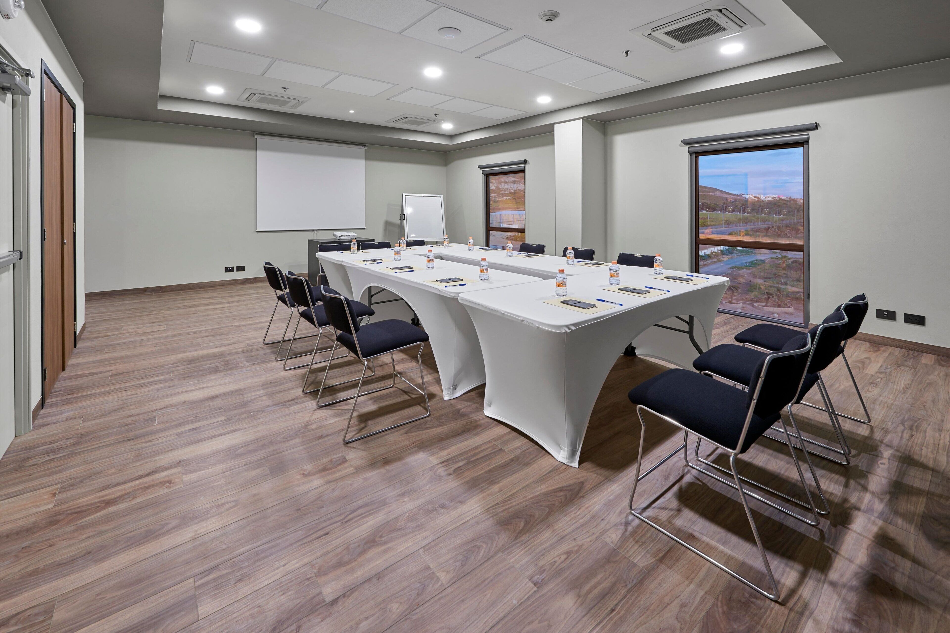 A conference room with a long table, chairs, a projector screen, and a view of a landscape through a large window.