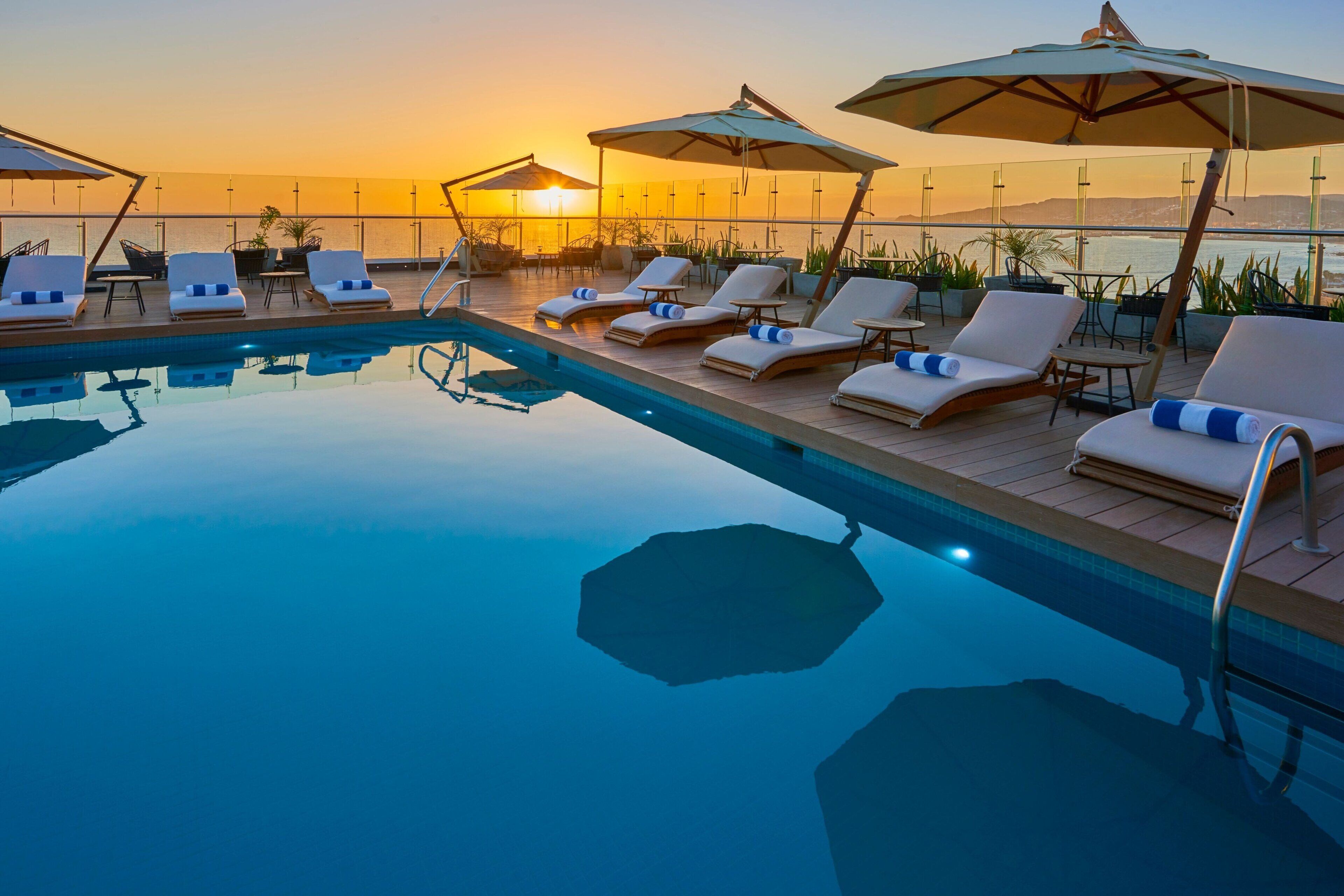 Zona junto a la piscina con sillones de descanso y sombrillas, con vista a un cuerpo de agua durante el atardecer.