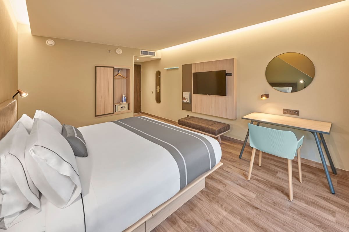 Ein modernes Hotelzimmer mit Bett, Nachttisch, Sessel, Schreibtisch, Spiegel und integriertem Fernseher.