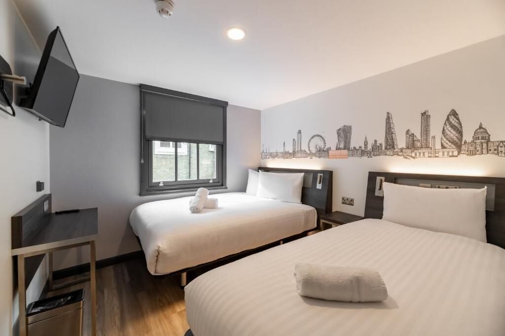 A hotel room with two beds, a TV, a window, and a cityscape mural on the wall.