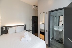 A hotel room with a large bed, a bedside table, a mirror, a bathroom with a sink and toilet, and a door leading to another room. - Easyhotel London Victoria (London)