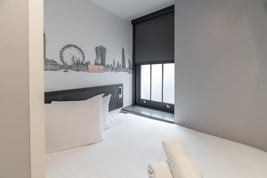 Habitación de hotel con cama, almohadas blancas, ventana con vistas y un mural en la pared que representa un horizonte urbano.