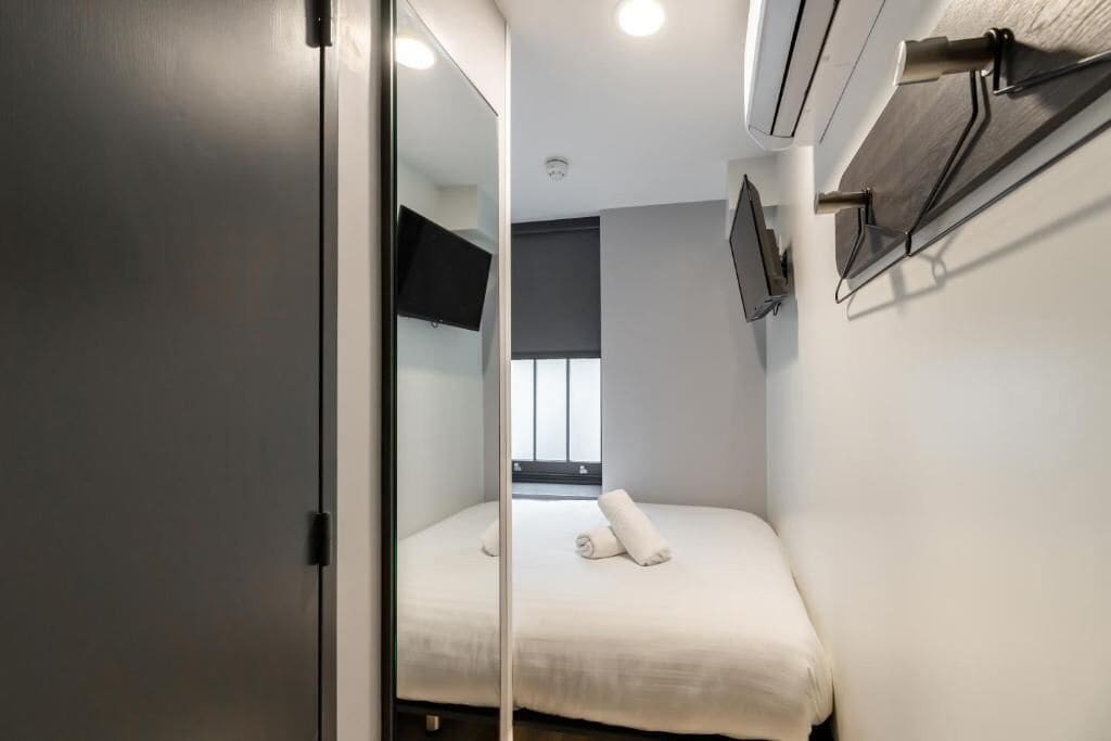 A narrow hotel room with a bed, a flat-screen TV mounted on the wall, and a window with blinds.