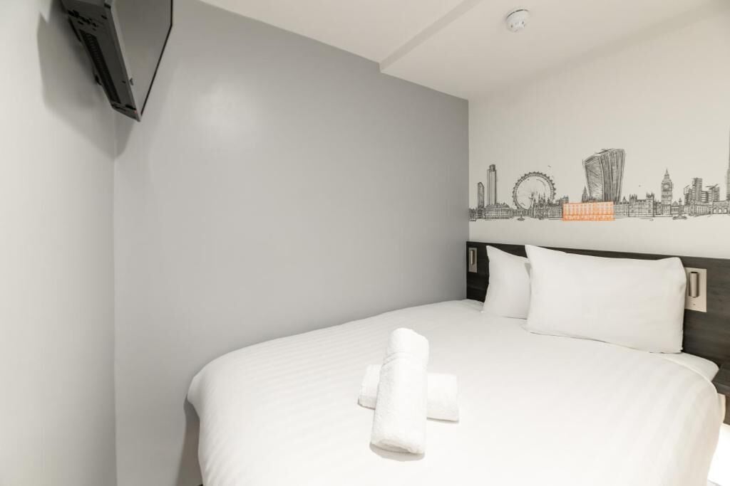 Habitación de hotel con cama, ropa de cama blanca, un mural con vista al horizonte de la ciudad y un televisor de pantalla plana montado en la pared.