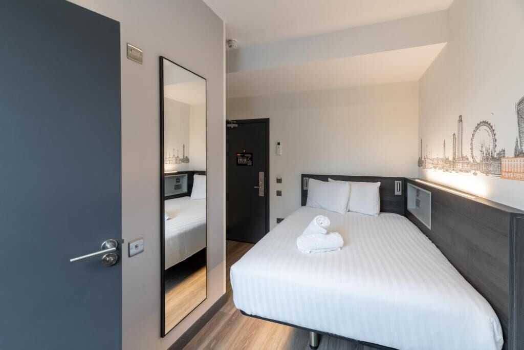 A hotel room with a bed, a large mirror, a door, and a wall with a cityscape mural.