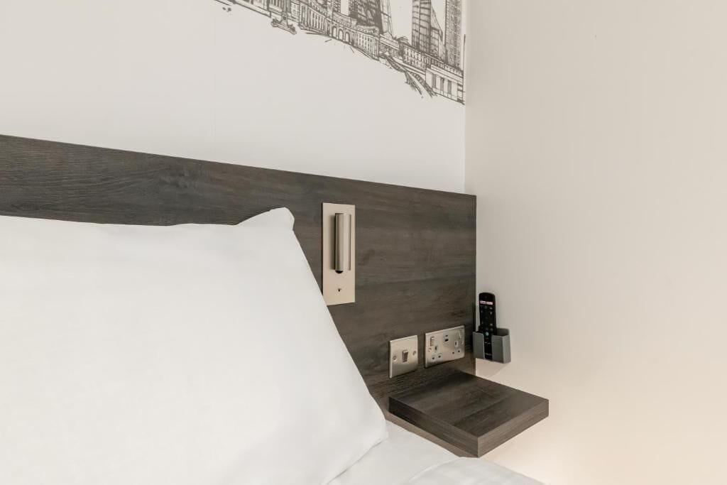 Quarto de hotel moderno com cabeceira de madeira, travesseiro branco, mesa de cabeceira com controle remoto e tomada na parede.