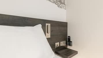 Een moderne hotelkamer met een houten hoofdbord, een wit kussen, een nachtkastje met afstandsbediening en een wandcontactdoos.