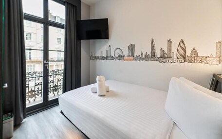 A hotel room with a bed, a cityscape wall art, a TV mounted on the wall, and a balcony with a view of buildings.