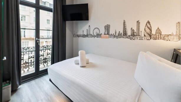 A hotel room with a bed, a cityscape wall art, a TV mounted on the wall, and a balcony with a view of buildings. - Easyhotel London Victoria (London)