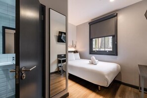 A modern hotel room with a bed, a desk, a chair, a flat-screen TV, and a window with a view of buildings. - Easyhotel London Victoria (London)
