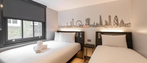 A hotel room with two beds, a cityscape wall art, a window with blinds, and a small bedside table.