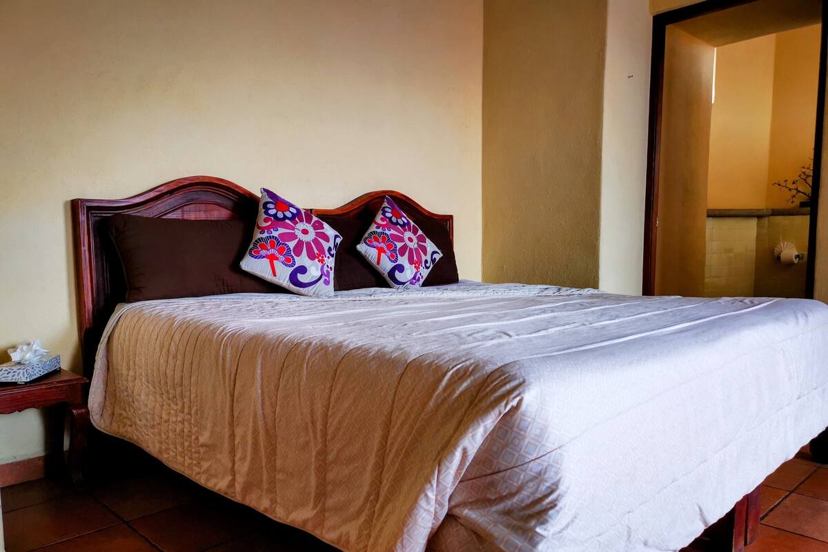 Una cama bien hecha con cubrecama acolchado, dos almohadas decorativas, un cabecero de madera y una mesita de noche con una caja de pañuelos.