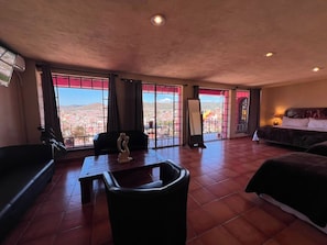 Habitación amplia con una cama grande, un sofá, una mesa y una silla. Se ve un paisaje urbano a través de grandes ventanales.